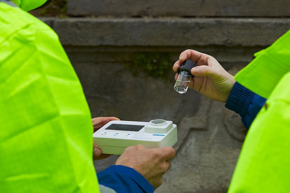 Operatori Nepta in abbigliamento ad alta visibilità effettuano un test della qualità dell'acqua utilizzando un fotometro digitale portatile e una provetta di campionamento in un sito esterno.