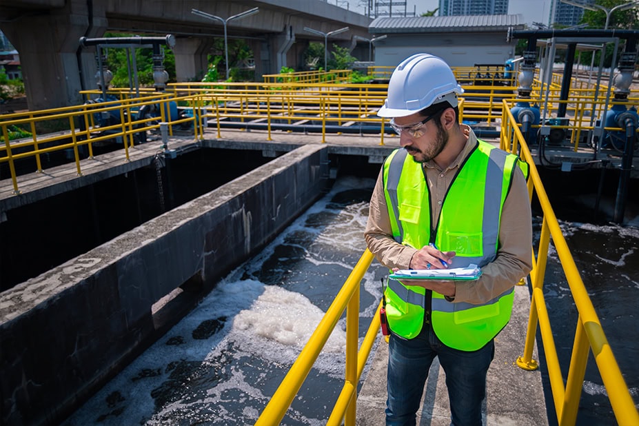 Ingegnere o tecnico con elmetto bianco e gilet ad alta visibilità esegue un'ispezione su un impianto di trattamento o depurazione delle acque, prendendo appunti su un tablet. L'immagine simboleggia l'applicazione del know-how in digitalizzazione e innovazione del Gruppo Italgas nello sviluppo di una rete idrica efficiente per Nepta