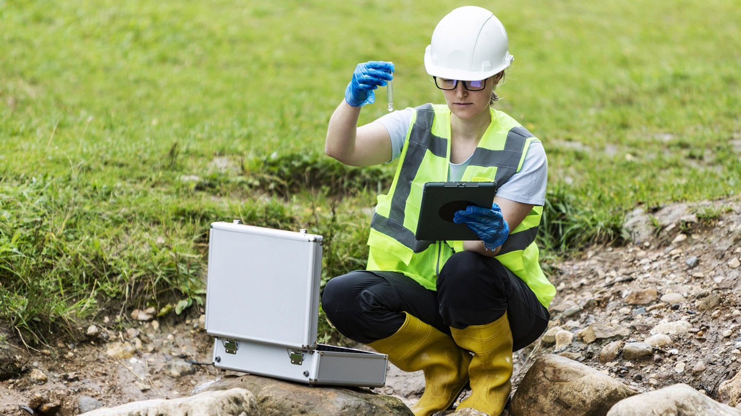Tecnica di laboratorio Nepta con casco bianco, giubbotto giallo ad alta visibilità e guanti blu preleva un campione d'acqua con provetta in ambiente esterno verde, tablet e valigetta aperta accanto.