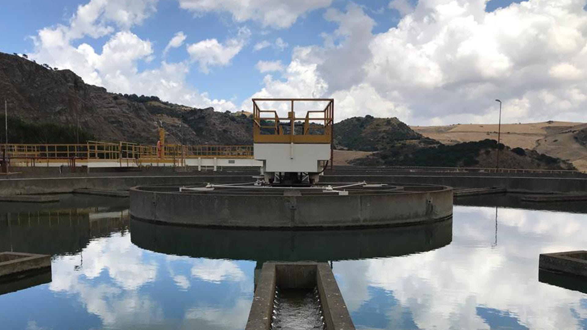Immagine panoramica di un impianto di trattamento, con un decantatore circolare e un cielo nuvoloso che si riflette sull'acqua. L'immagine rappresenta gli impianti gestiti da Siciliacque, gestore del servizio idrico di sovrambito per la Regione Sicilia dal 2004 (Gruppo Nepta/Italgas).