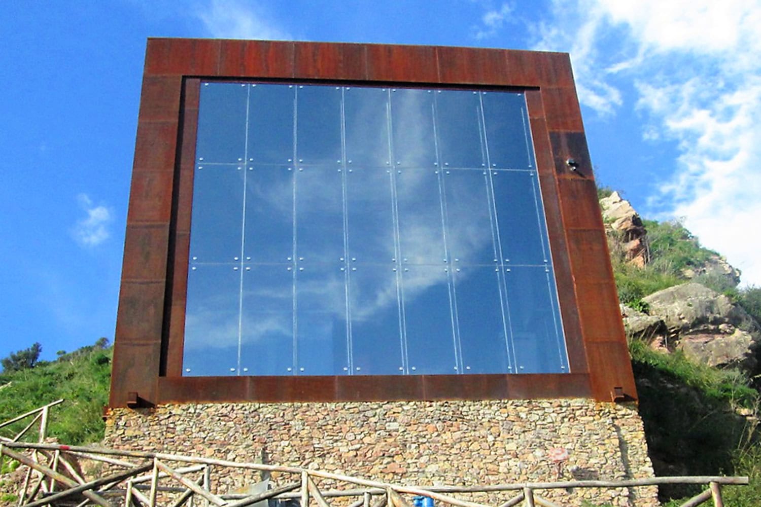 Edificio moderno in acciaio Corten arrugginito e pietra, con una grande facciata vetrata che riflette il cielo blu, situato su una collina. L'immagine simboleggia la trasformazione e l'innovazione tecnologica che Siciliacque (Gruppo Nepta/Italgas) sta applicando alle infrastrutture idriche essenziali per la sicurezza idrica della Sicilia.