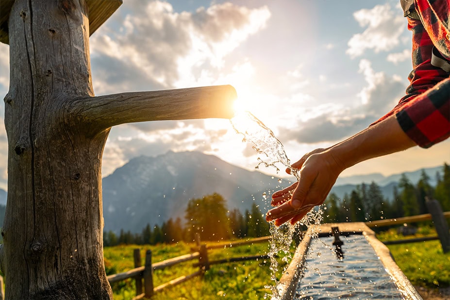 Primo piano di una mano che raccoglie acqua fresca e limpida che sgorga da una fontana rustica in legno, con uno sfondo montano illuminato dal sole. L'immagine simboleggia la purezza dell'acqua dalla fonte e invita a scoprire l'intero percorso dell'acqua gestito dal Gruppo Nepta/Italgas.