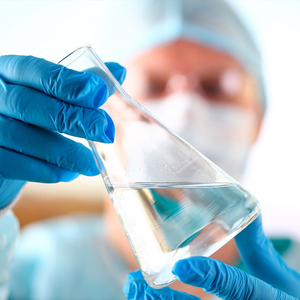 Primo piano di un tecnico di laboratorio (indossando mascherina, cuffia e guanti blu) che esamina un'ampolla o beuta contenente acqua limpida. L'immagine simboleggia il rigoroso controllo qualità e il monitoraggio costante dell'acqua da parte di Nepta/Gruppo Italgas lungo tutta la rete idrica.