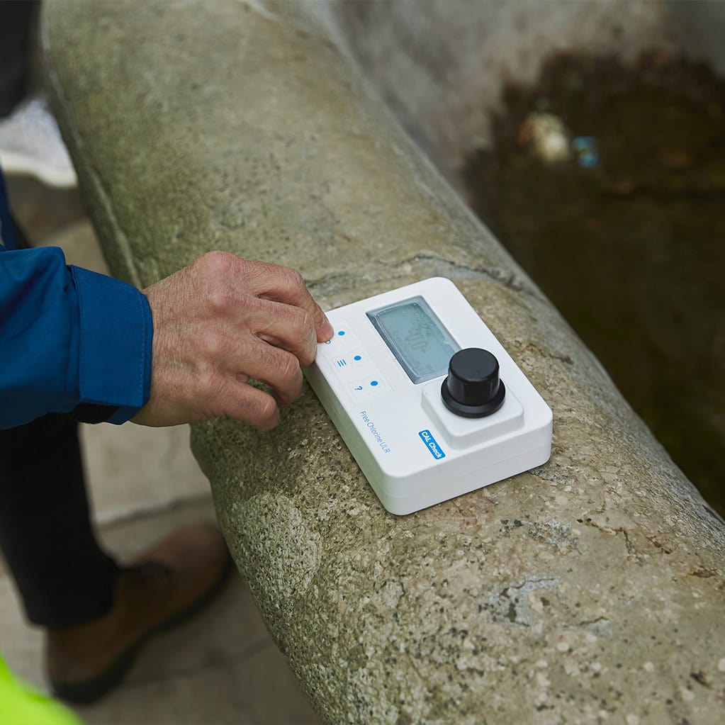 Primo piano di una persona che utilizza un fotometro portatile o un altro strumento di misurazione della qualità dell'acqua su un bordo in pietra, probabilmente per analizzare un campione prelevato da una fontana o una vasca. L'immagine simboleggia i controlli sul campo e il trattamen