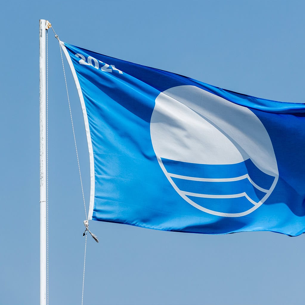Bandiera Blu sventola contro un cielo azzurro, simbolo di alta qualità delle acque di balneazione e del servizio idrico. L'immagine rappresenta il successo della depurazione delle acque gestita da Nepta/Gruppo Italgas (tramite Acqualatina) e la restituzione all'ambiente di una risorsa pulita.