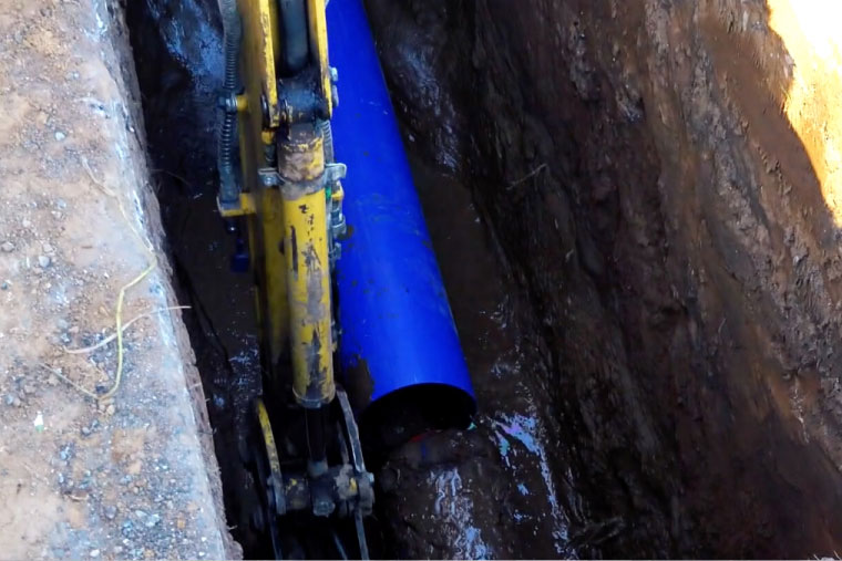 Dettaglio ravvicinato di uno scavo per la posa di una nuova tubazione idrica blu con l'utilizzo di una macchina operatrice. Simbolo degli interventi di Acqualatina S.p.A.