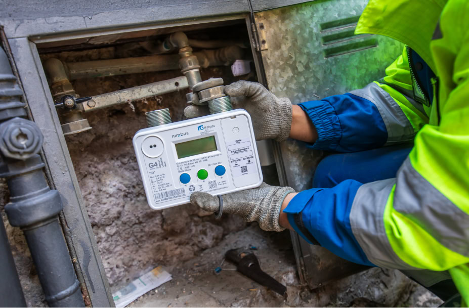 Un operaio installa un contatore del gas Nimbus di Italgas in una nicchia, tenendolo con guanti da lavoro. Il contatore è di colore bianco con un display digitale.