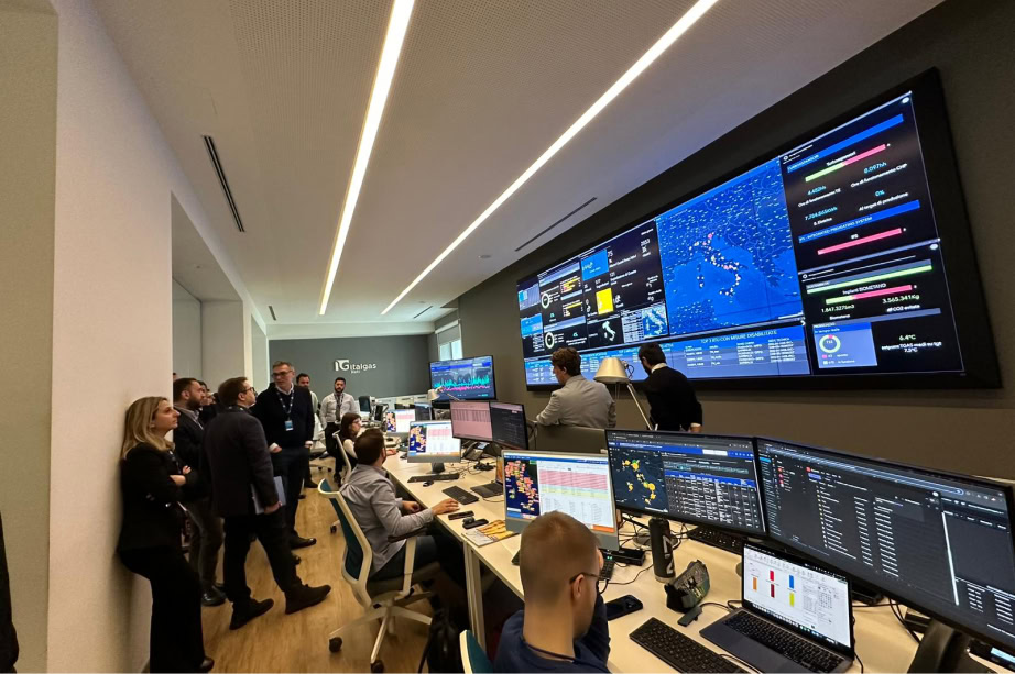 Sala di controllo Italgas: persone in piedi e sedute monitorano schermi con dati e grafici.