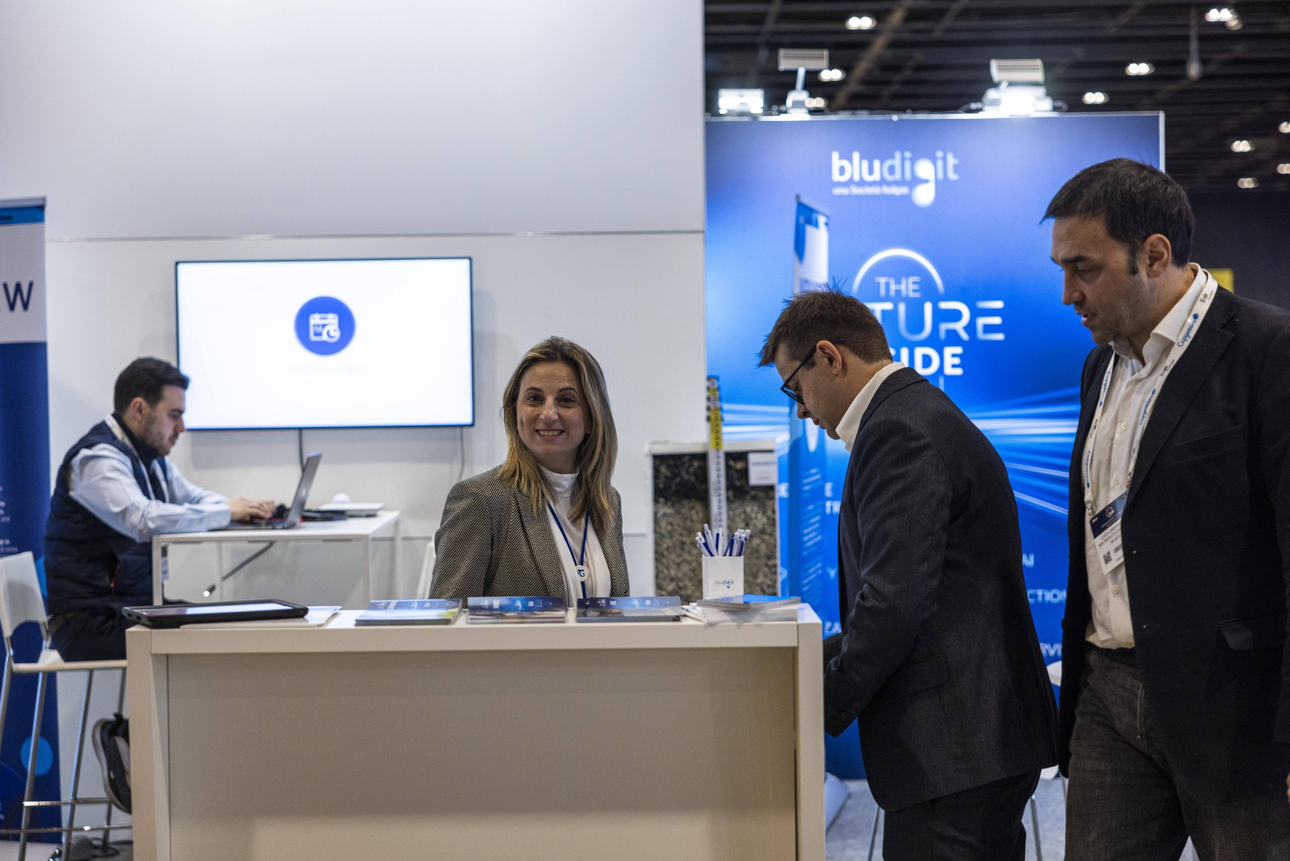 A business event with people at a reception desk. A woman smiles while two men read promotional material. In the background, a man works on a computer.