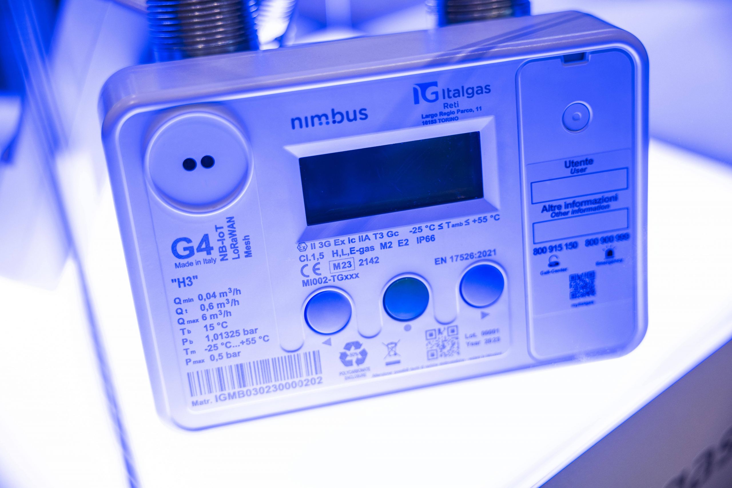 Blue-lit image of an Italgas brand nimbus gas meter with display and buttons, technical labels visible.