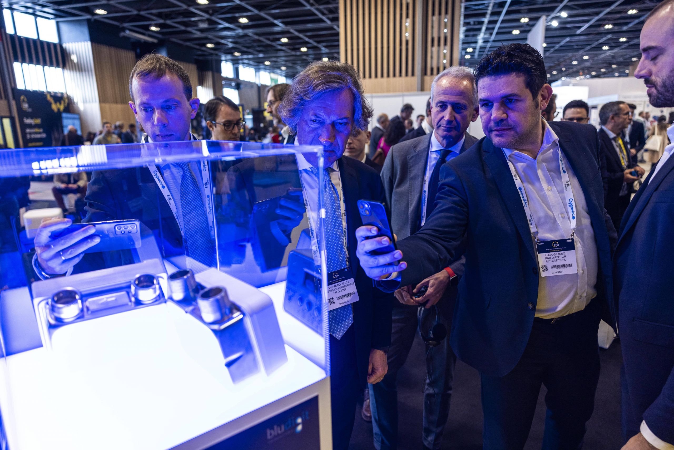 Some men in business attire observe and photograph with their phones the Nimbus in a blue-lit case during a professional event at a trade fair.