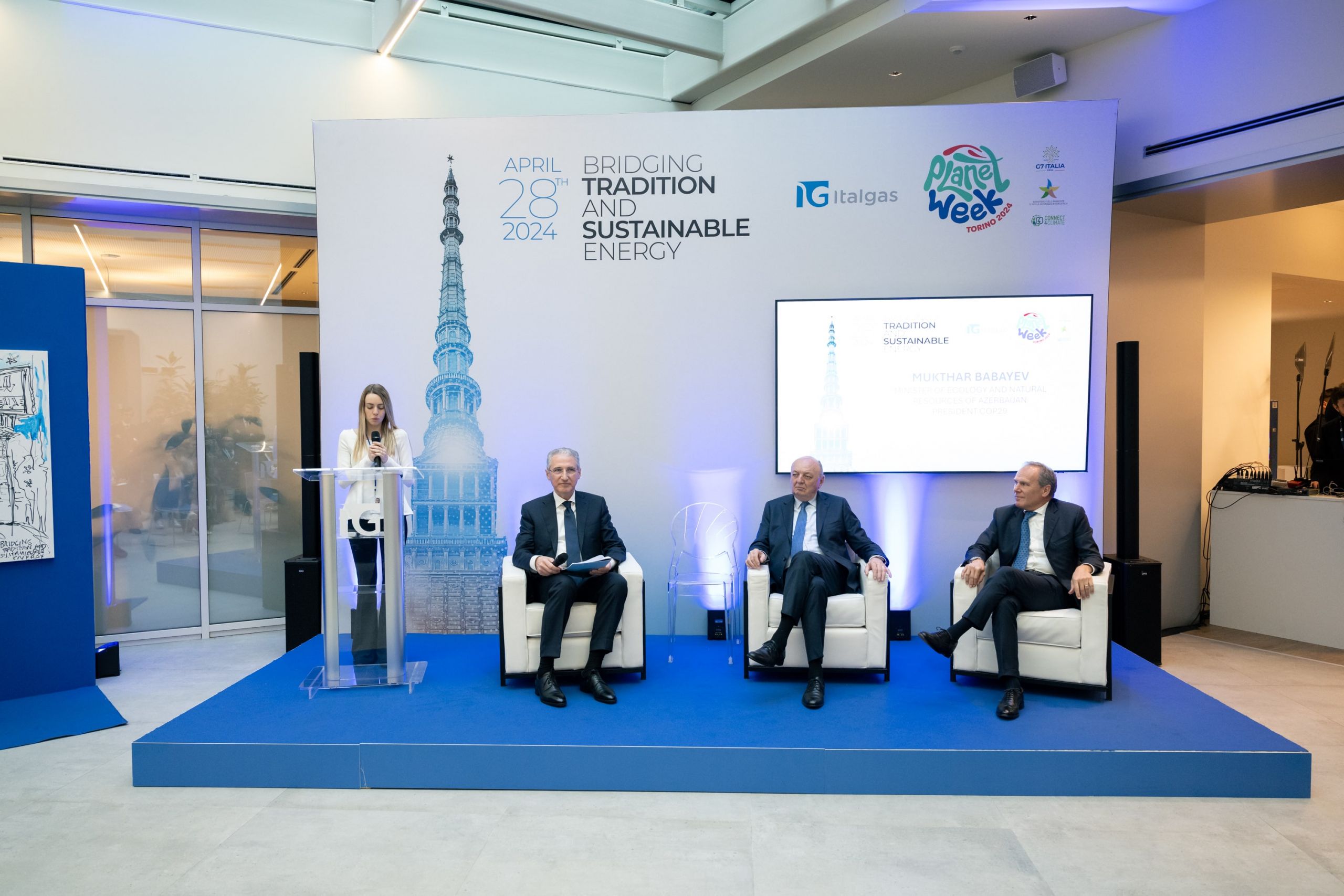 Woman presenting at Italgas Planet Week event with two seated men against a backdrop with an image of a tower.