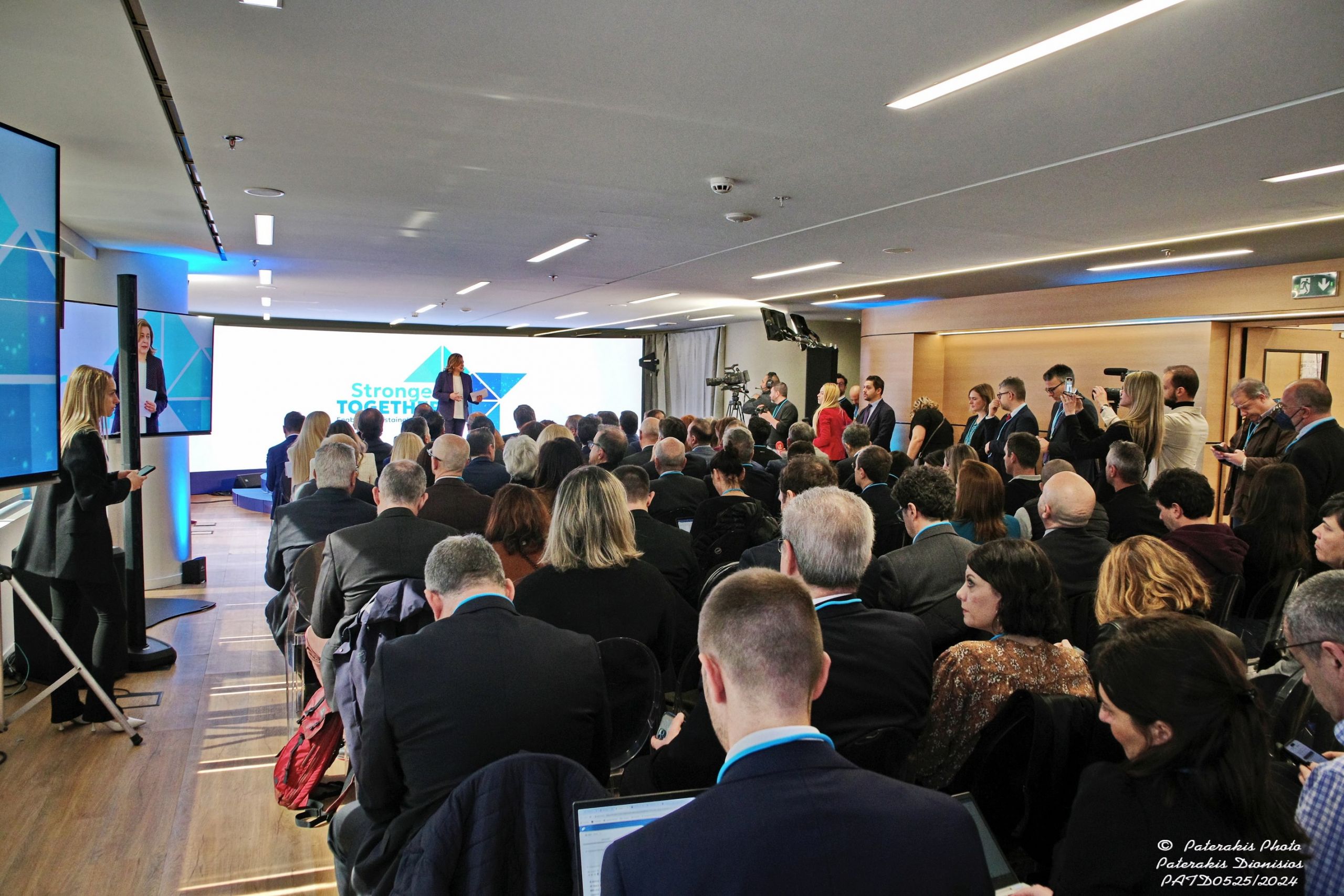 Conference room with attendees focused on a speaker beside a 'Stronger Together' slide.