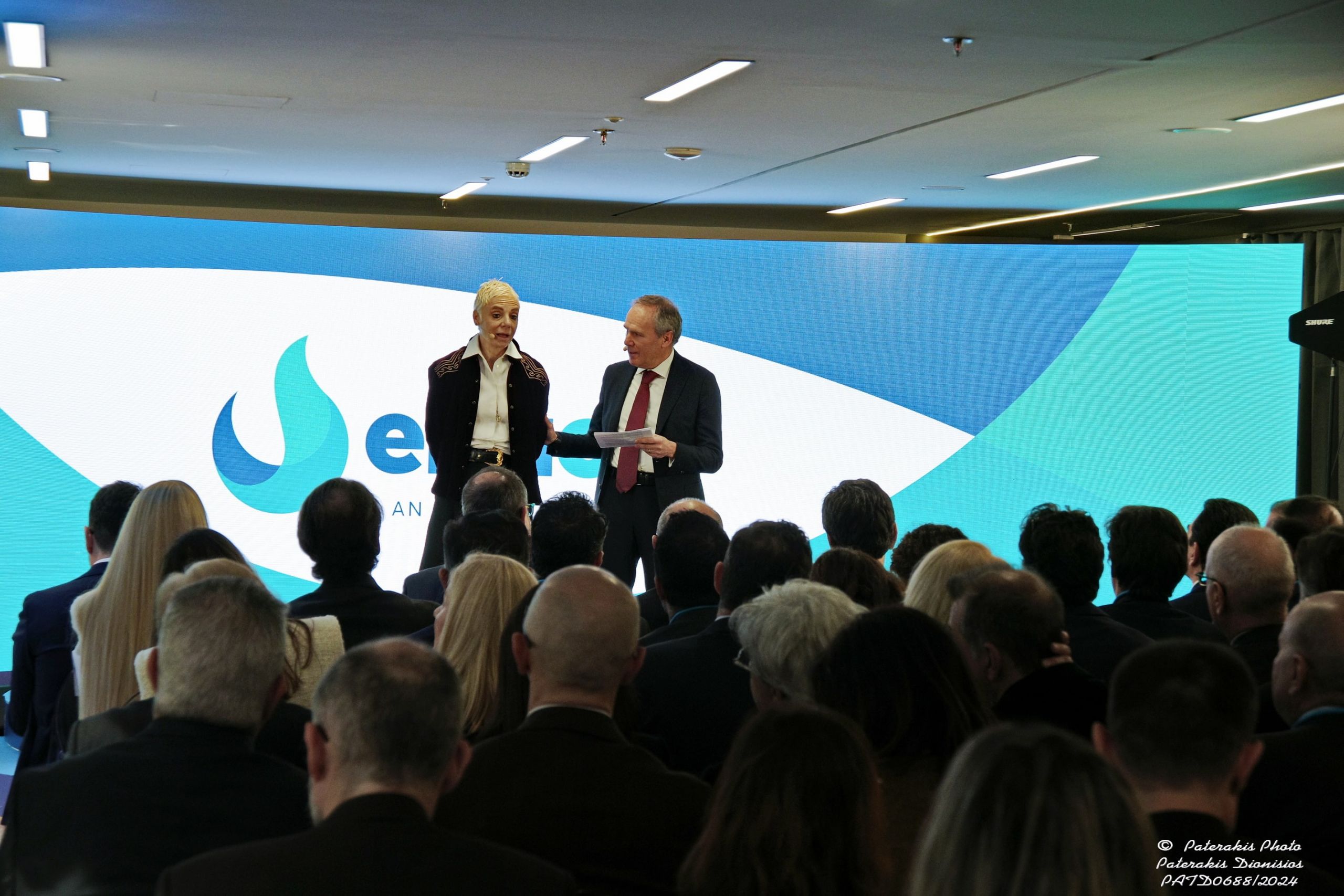 Two people presenting at a corporate event with audience in foreground and the screening of the Enaon logo in the background.