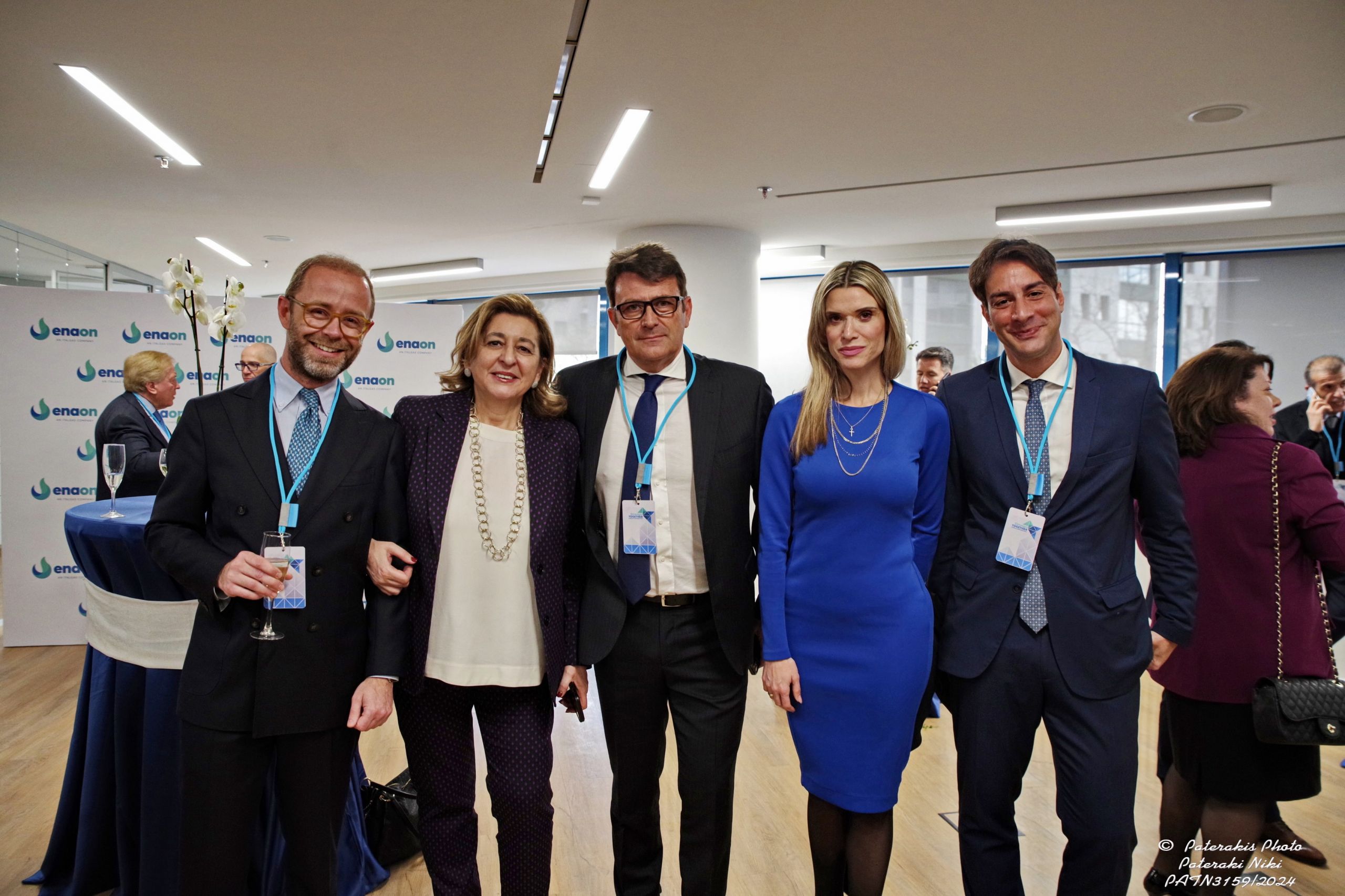 Five professionals in business attire pose smiling at a corporate event with logo banners in the background.