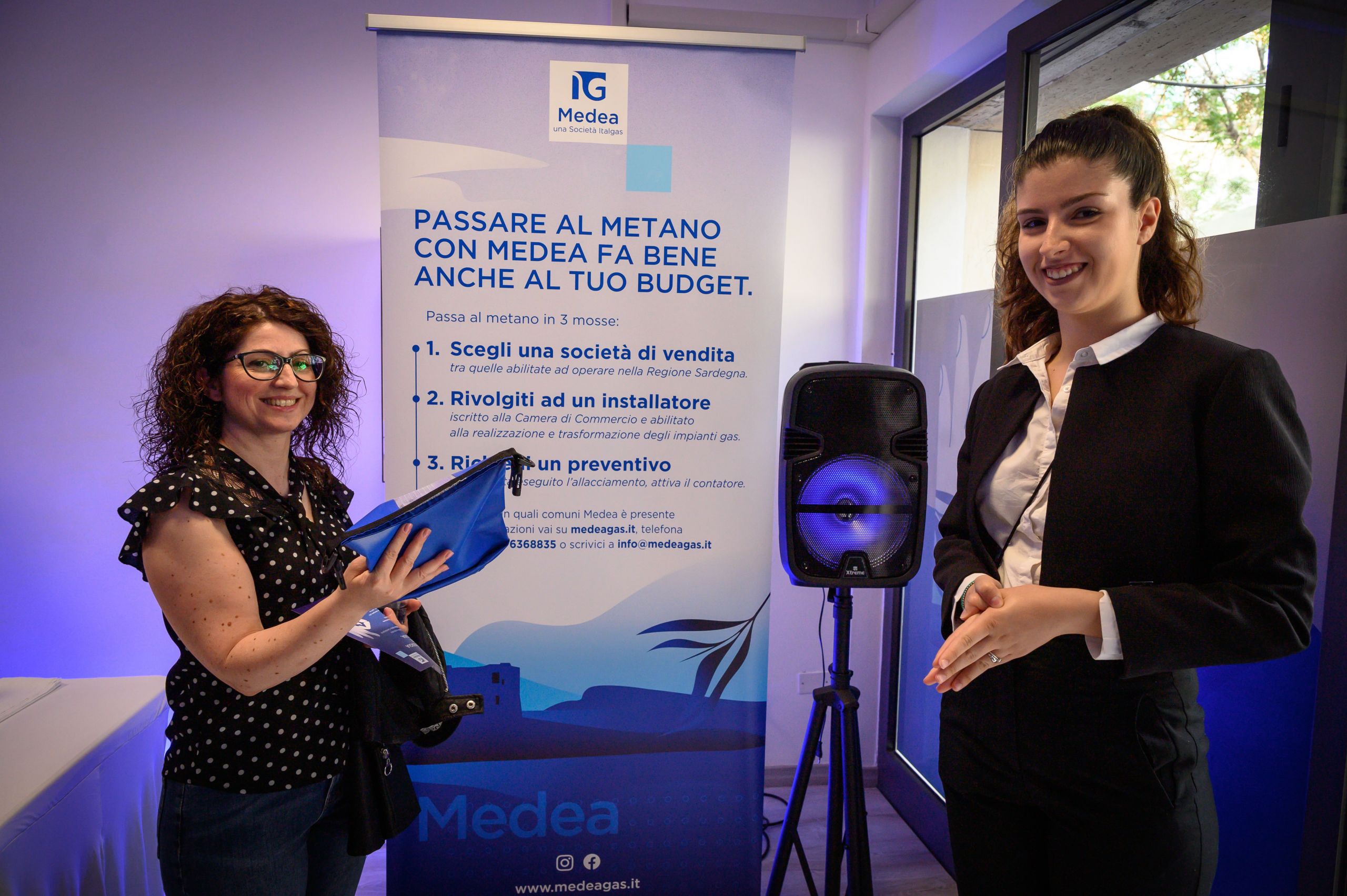 Two smiling women at Medea Italgas infopoint with informational banner and speaker.