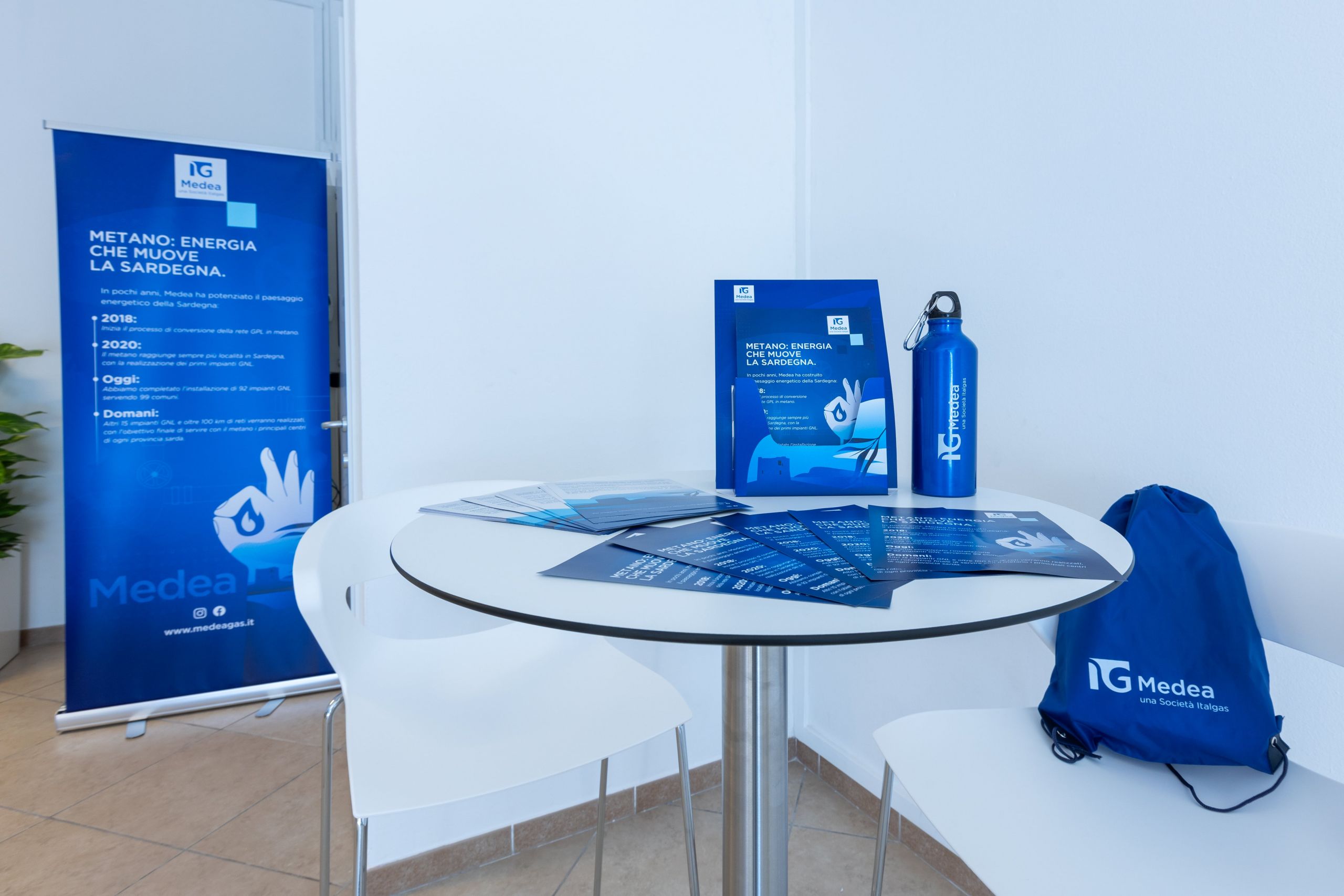 Infopoint setup with Medea Italgas branding on materials like flyers, a bottle, and a backpack on a white table.