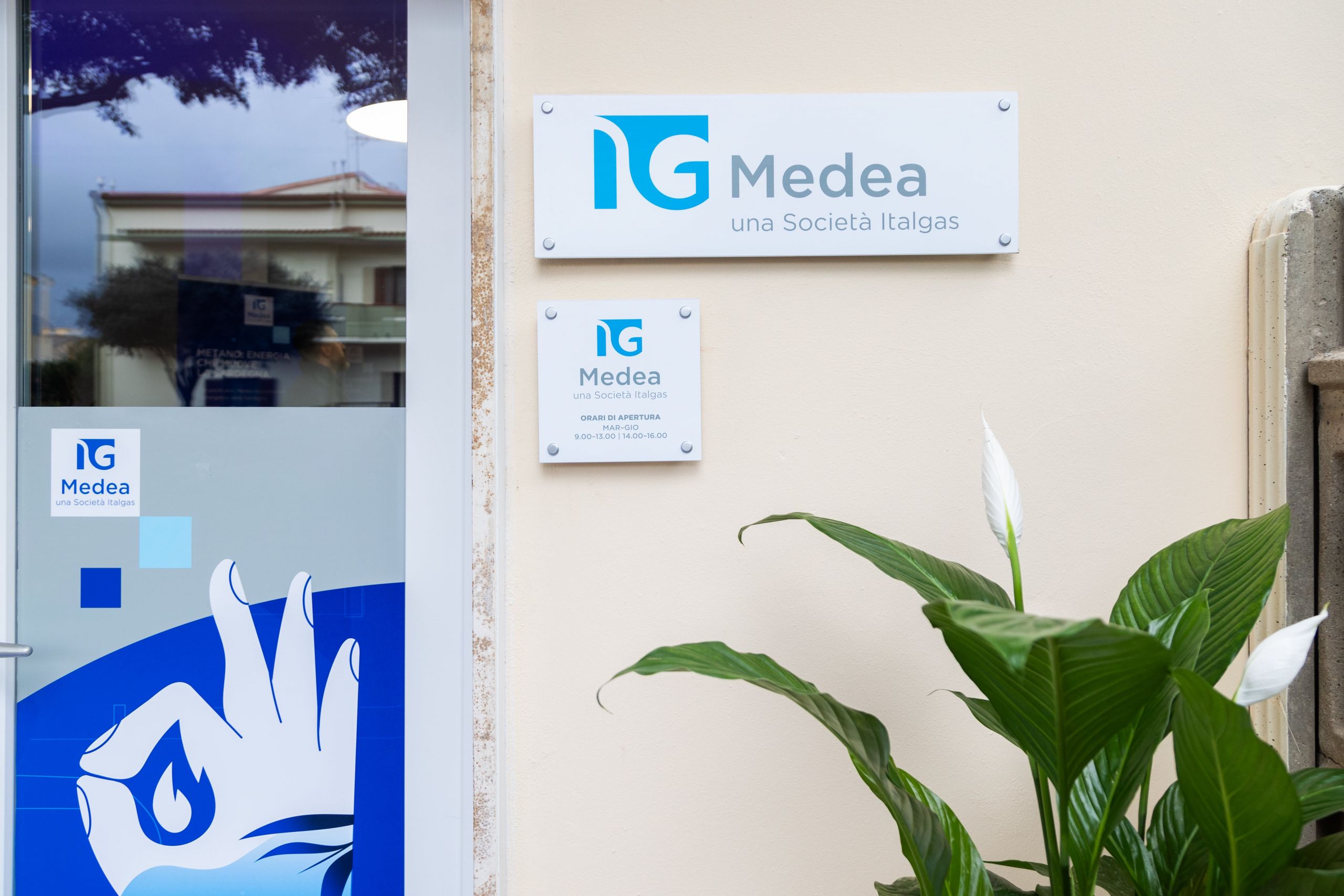 Signs for Medea, a Società Italgas brand, on wall beside a window with reflections and plants in foreground.
