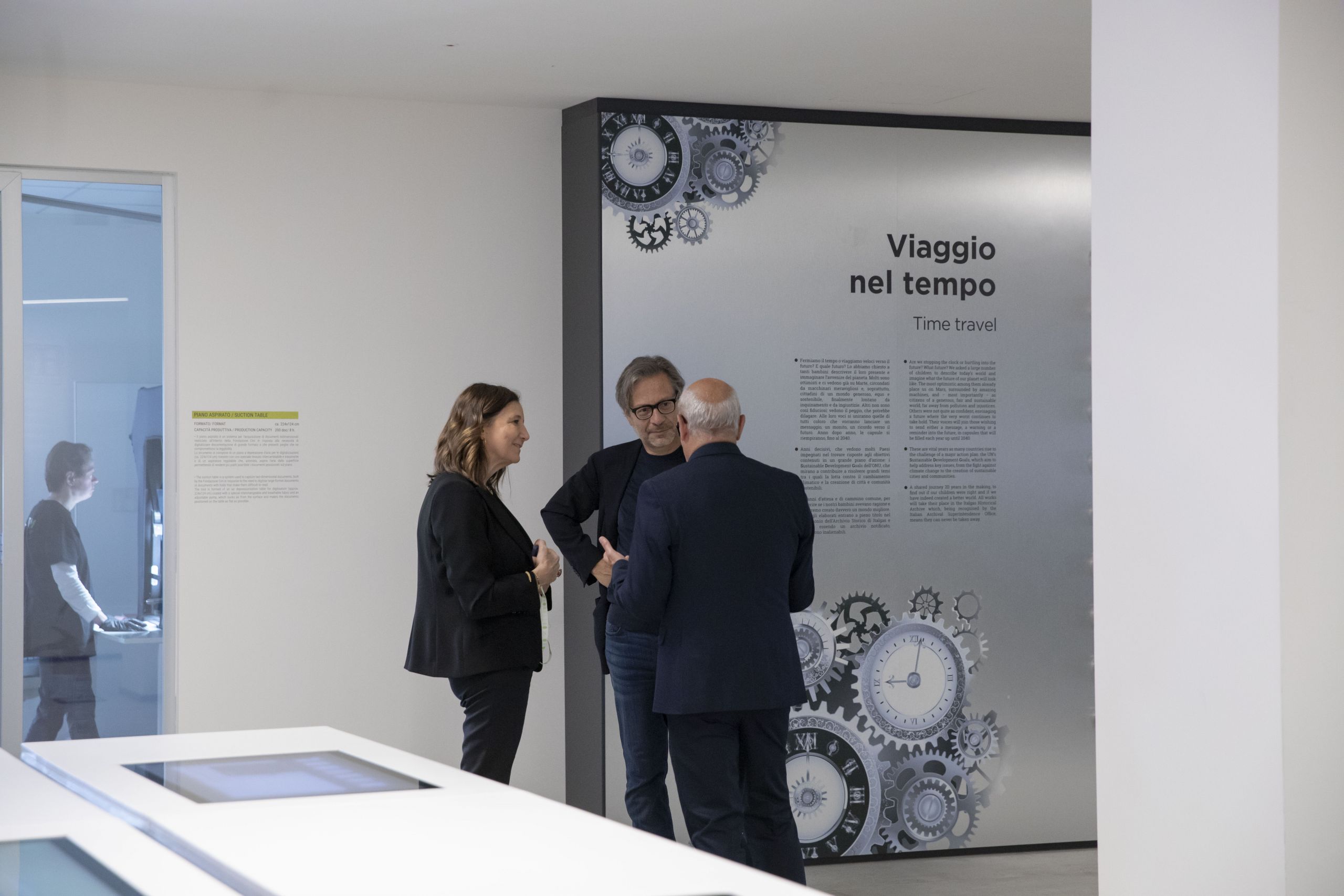 Three people conversing by an exhibition titled 'Viaggio nel tempo/Time travel,' with cogwheel graphics.