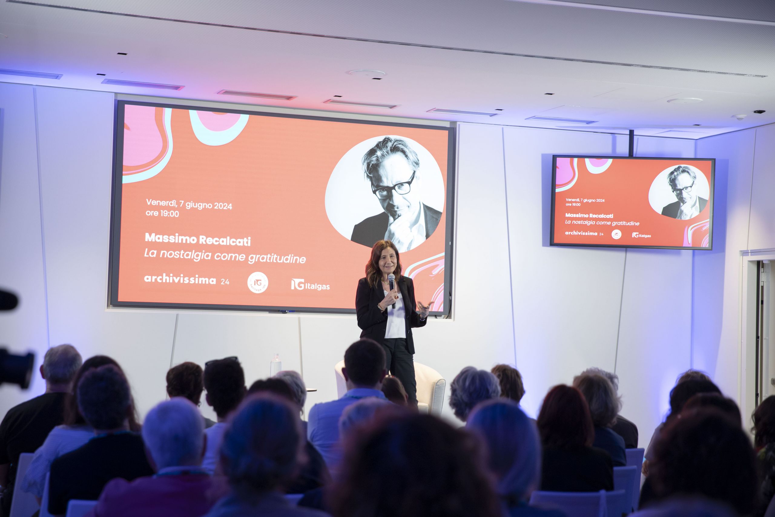 Woman presenting at Heritage lab event with audience and a large screen displaying event details.