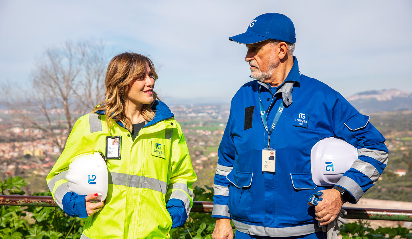 Due dipendenti Italgas un uomo e una donna in divise da lavoro e caschi protettivi conversano all'aperto.