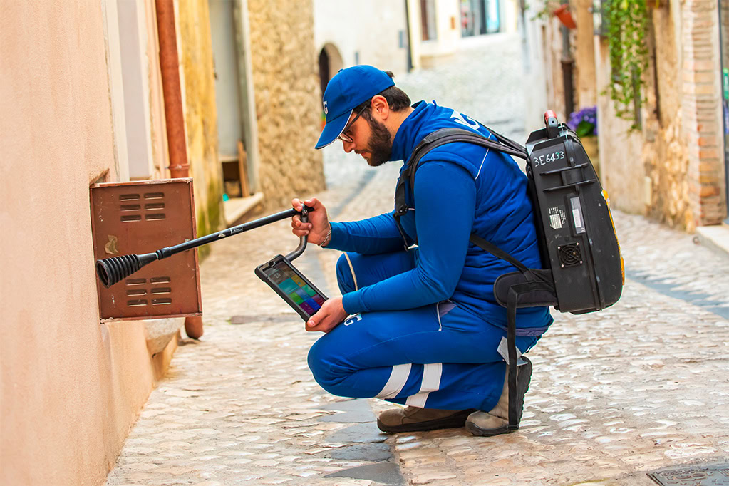 Tecnico Italgas accovacciato utilizza la lancia dello zaino Picarro per controllare dispersioni di gas all'interno di un vano contatore. In mano ha un tablet per controllare i dati. 