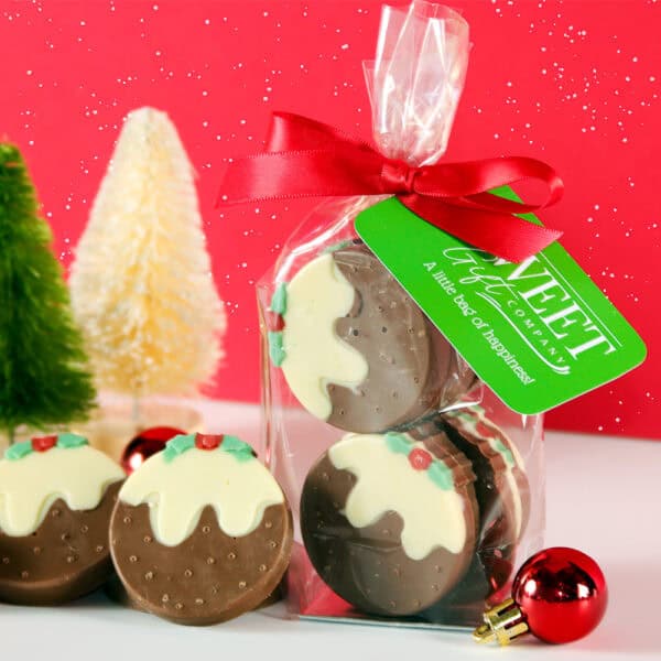 A clear bag of Mini Chocolate Christmas Puddings, adorned with a red ribbon and green tag, positioned beside miniature Christmas trees and ornaments on a festive backdrop.