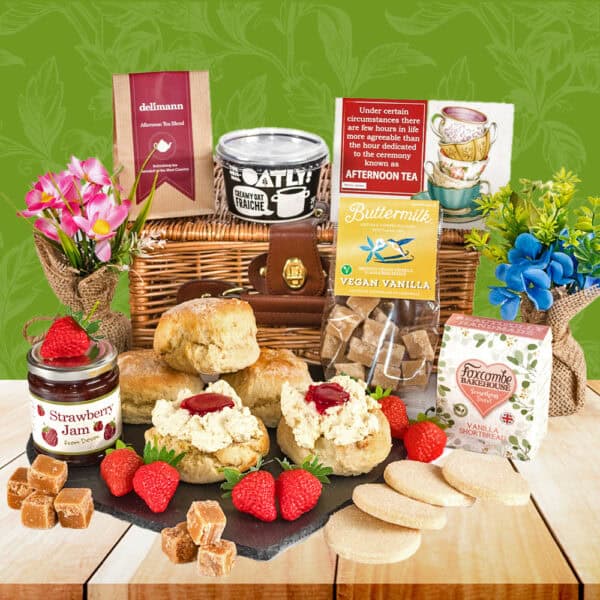 The Vegan Cookies & Cream Tea Hamper includes a wicker basket filled with scones, oat milk, vegan sweets, strawberry jam, vegan clotted cream, shortbread, fudge, strawberries, and two floral mugs.