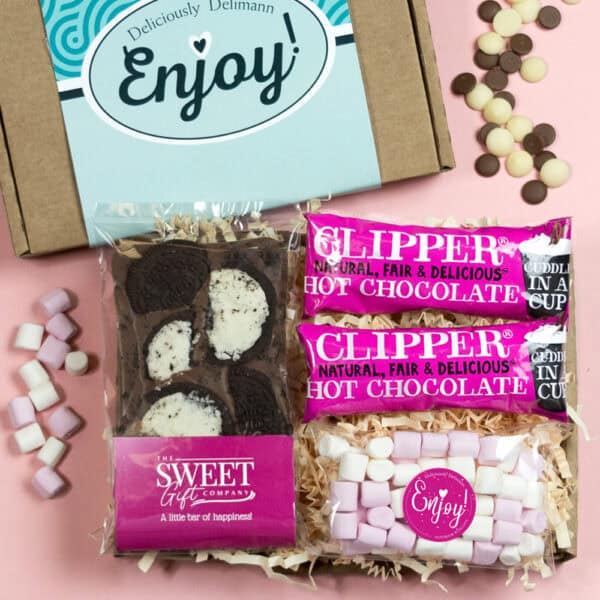 The Little Hot Chocolate Letterbox Gift is a charming box marked "Enjoy!" with two Clipper hot chocolate sachets, a cookies chocolate bar, and mini marshmallows, set on a pink background with chocolate chips and marshmallows.