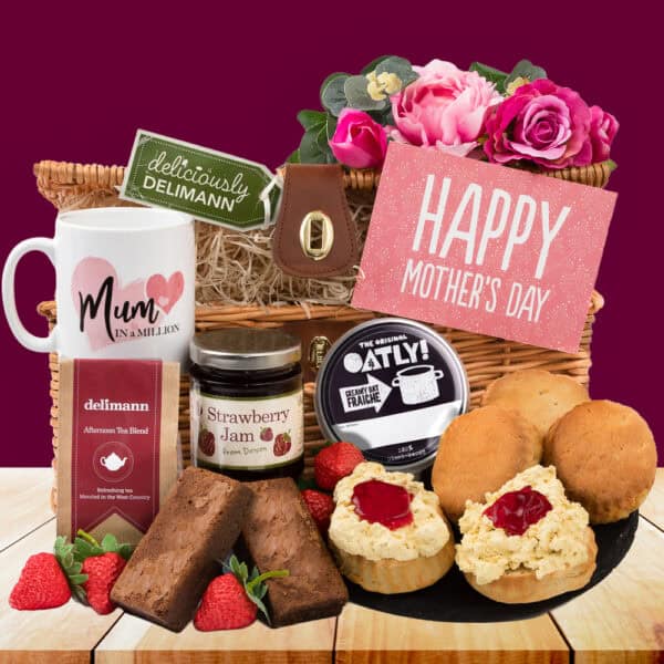 The Mother’s Day Vegan Cream Tea Gift features a “Mum in a Million” mug, tea, brownies, strawberry jam, Oatly cream, scones with jam and cream, a “Happy Mother’s Day” sign, pink flowers, and strawberries on a wooden surface.