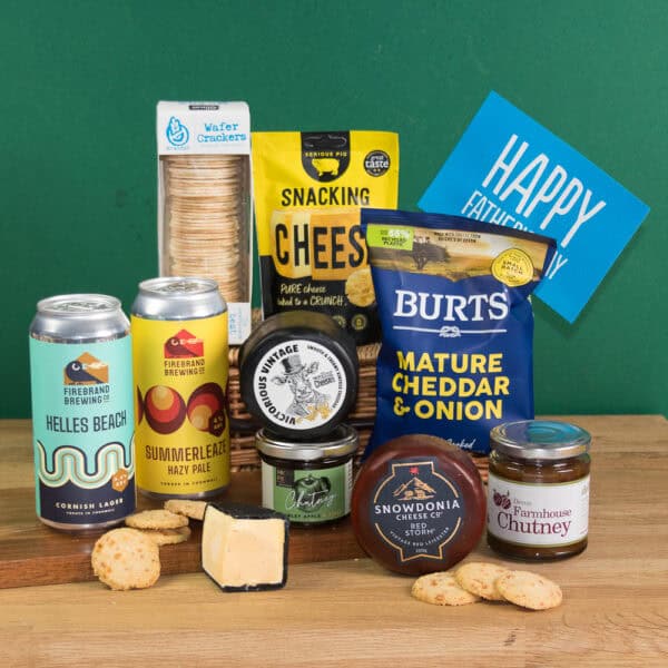 A selection of snacks and drinks, including cheese, biscuits, chutney, cheese crisps, and Dad's Gluten Free Cheese & Cornish Beer Gift with a “Happy Father’s Day” card, displayed on a wooden surface against a green background.