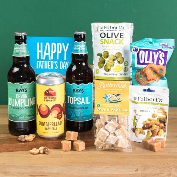 Surprise Dad with the Dad's Vegan Devon & Cornish Beer Box Gift: two Bays beers, Firebrand Summerleaze Hazy Pale Ale, fudge, olives, nuts, pretzels, and a blue "Happy Father's Day" card—all packed together on a wooden surface.