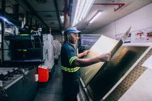 Printer Thabo Mnisi inspects a sheet of 729 printed metal crowns, prior to varnishing, pressing out, and the final stage of lining.