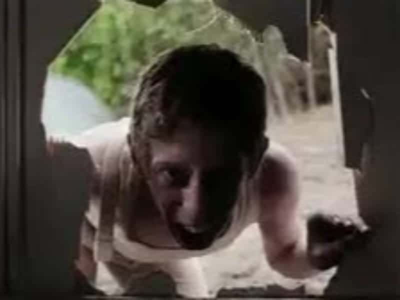 A person leans through a large broken hole in a door, looking inside with an intense expression—just like a scene from classic horror films. Trees and grass are visible outside.