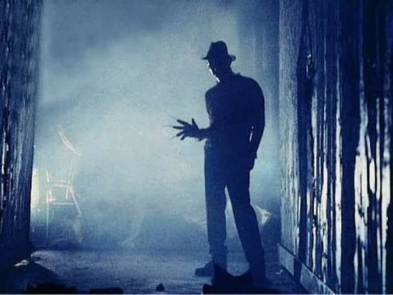 A silhouette of a person wearing a hat stands in a dark, narrow hallway, evoking classic Horror Movies with an outstretched hand tipped with long, sharp blades.