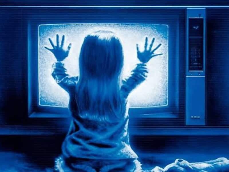 A young child sits in front of an old television displaying static, echoing classic horror movies, touching the screen with both hands in a blue-tinted room.