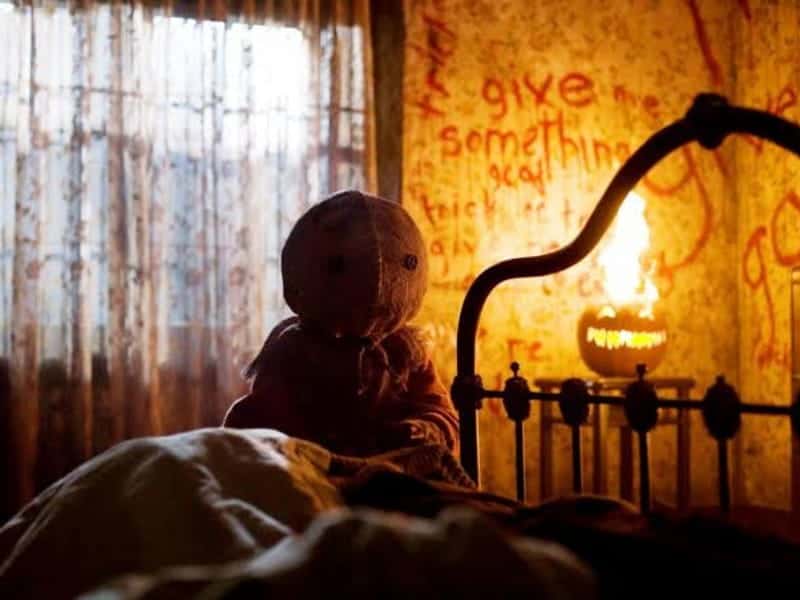 A fabric doll sits on a bed in a dim room. Red writing marks the wall, with a lit jack-o’-lantern—perfect for fans of Horror Movies during the 31 Days of fright.