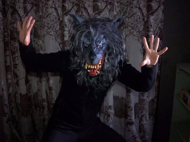 A person in a black outfit wearing a snarling werewolf mask poses with hands raised, ready for 31 Days of Horror Films, in front of a curtain with tree patterns.