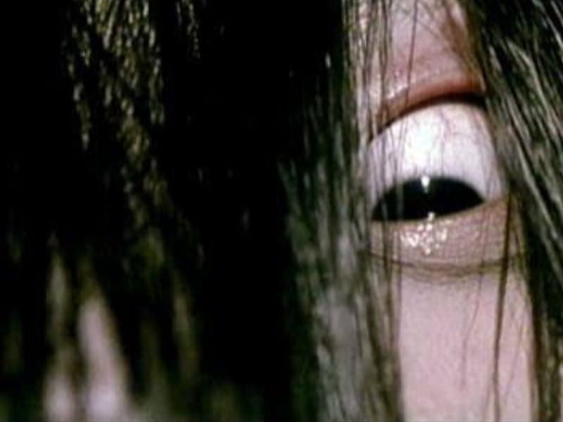 A close-up of a pale human eye partially obscured by long black hair, evoking the chilling atmosphere found in classic horror films.