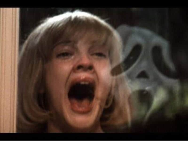 A woman with blonde hair screams in fear by a window, with a white ghost mask reflected behind her—a chilling homage to classic horror films.