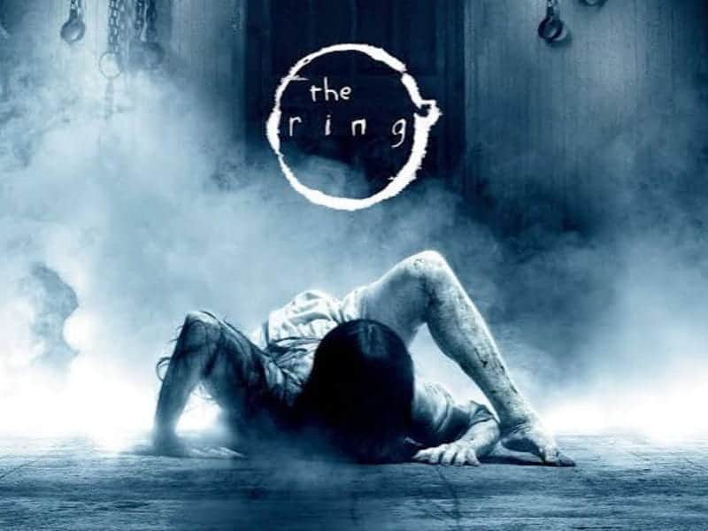 A figure with long dark hair crawls from smoky shadows. “The Ring” appears above—perfect for your 31 Days of Horror Movies binge. Chains dangle menacingly in the background.