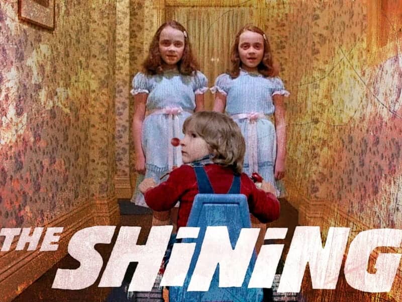 A boy on a tricycle faces two identical girls in blue dresses in a hallway—a chilling scene from one of the most iconic horror films, “The Shining.”.