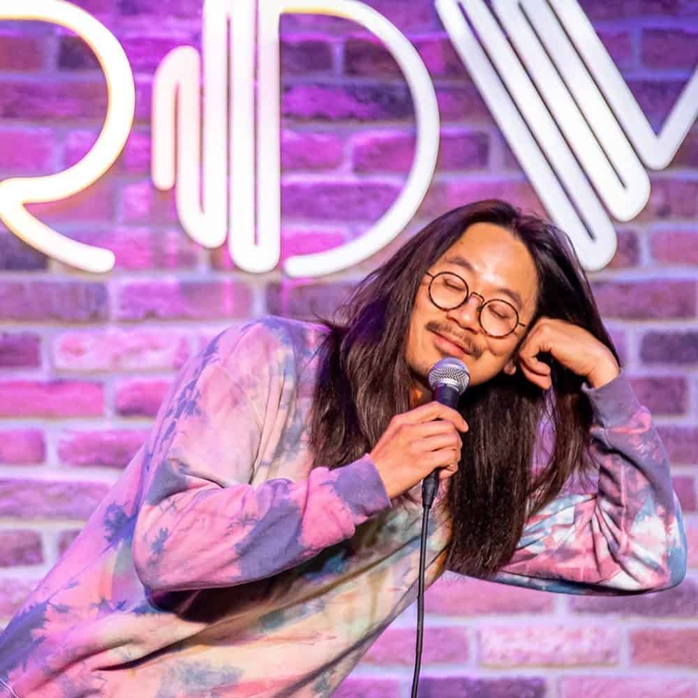 A person with long hair and glasses holds a microphone and leans on their hand, standing in front of a brick wall with a neon sign. Mark your calendars for May 16, 2024—this scene is part of your Must List!