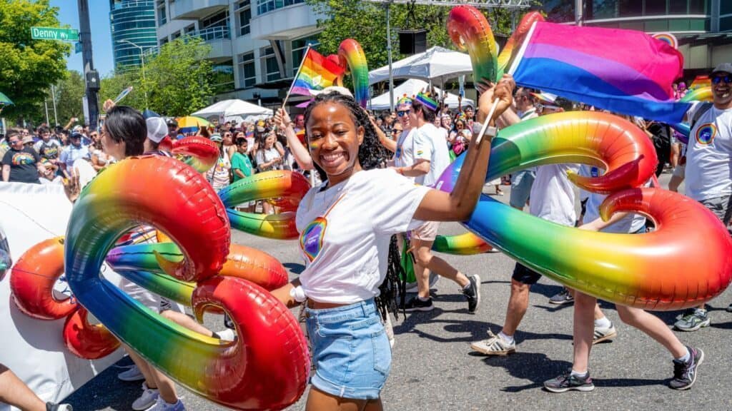 A group of people wearing rainbow-themed clothing and holding inflatable rainbow hearts participate in a pride parade under sunny conditions.