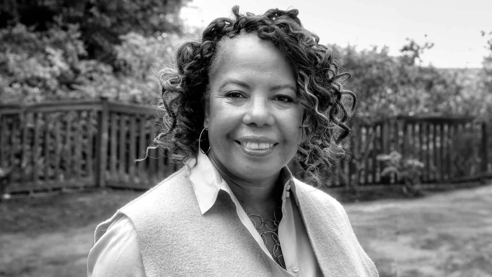 Lynne-Varner_1600x900 A trailblazing woman with curly hair smiles while standing outside in front of a wooden fence, captured beautifully in black and white.