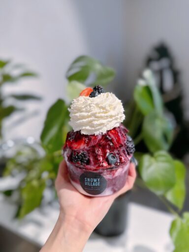Hand holding a dessert cup filled with shaved ice topped with fruit compote, fresh berries, and a generous swirl of whipped cream, with green plants blurred in the background—perfect for those Summer Vibes.