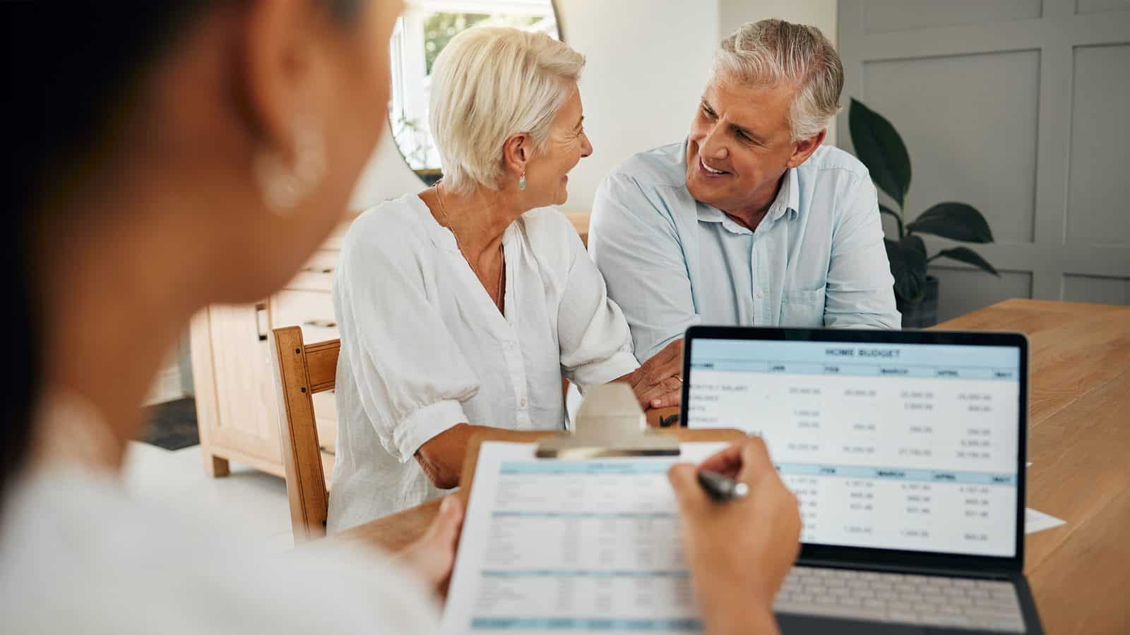FinancePlanningForRetirement_1600x900 A senior couple sits and smiles at each other while a professional, showcasing a sense of style, presents them with a financial document and a laptop displaying their retiring home budget spreadsheet on the table.
