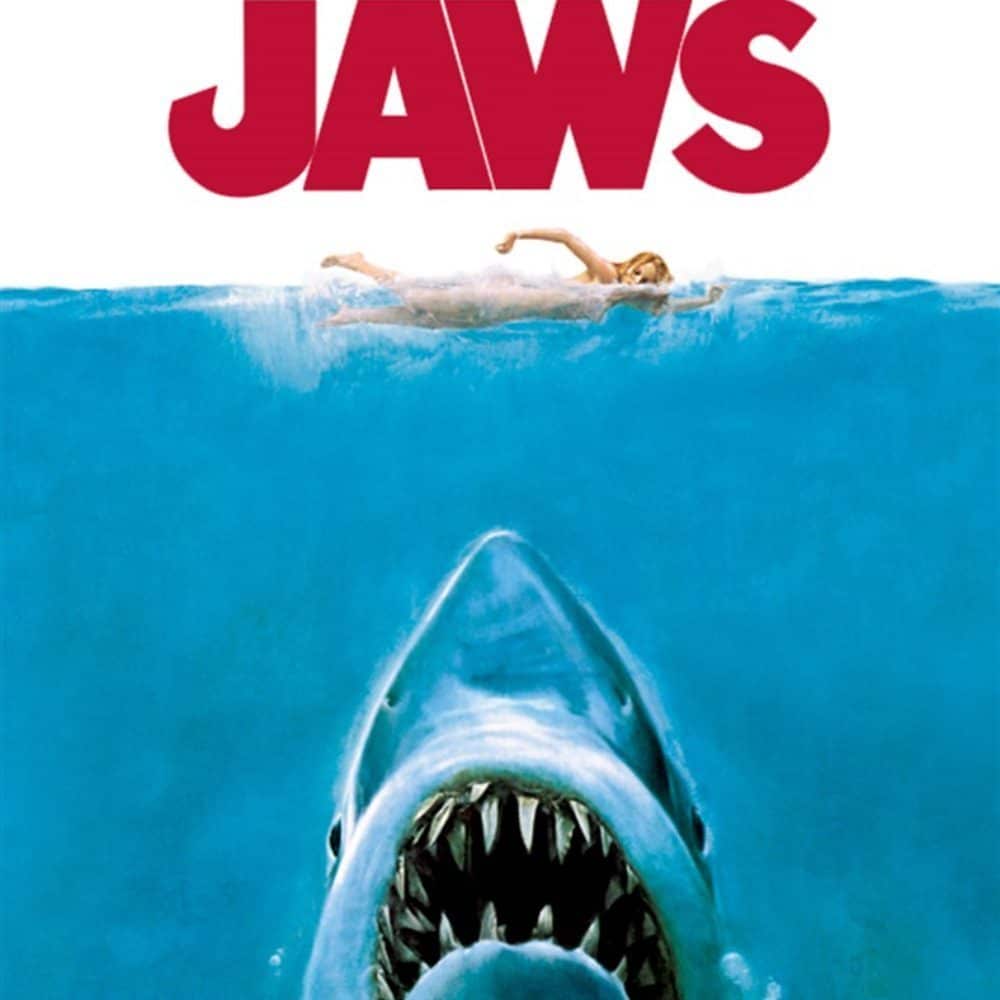 A swimmer is shown above water with a large, menacing shark emerging from below. The word "JAWS" is prominently displayed at the top in bold red letters—a must-list experience premiering Aug. 29.
