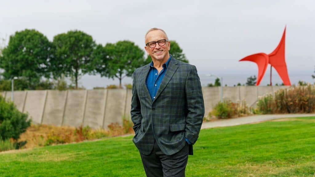 A man in a checkered blazer and glasses stands on a grassy area with trees, a concrete wall, and a red sculpture in the background.