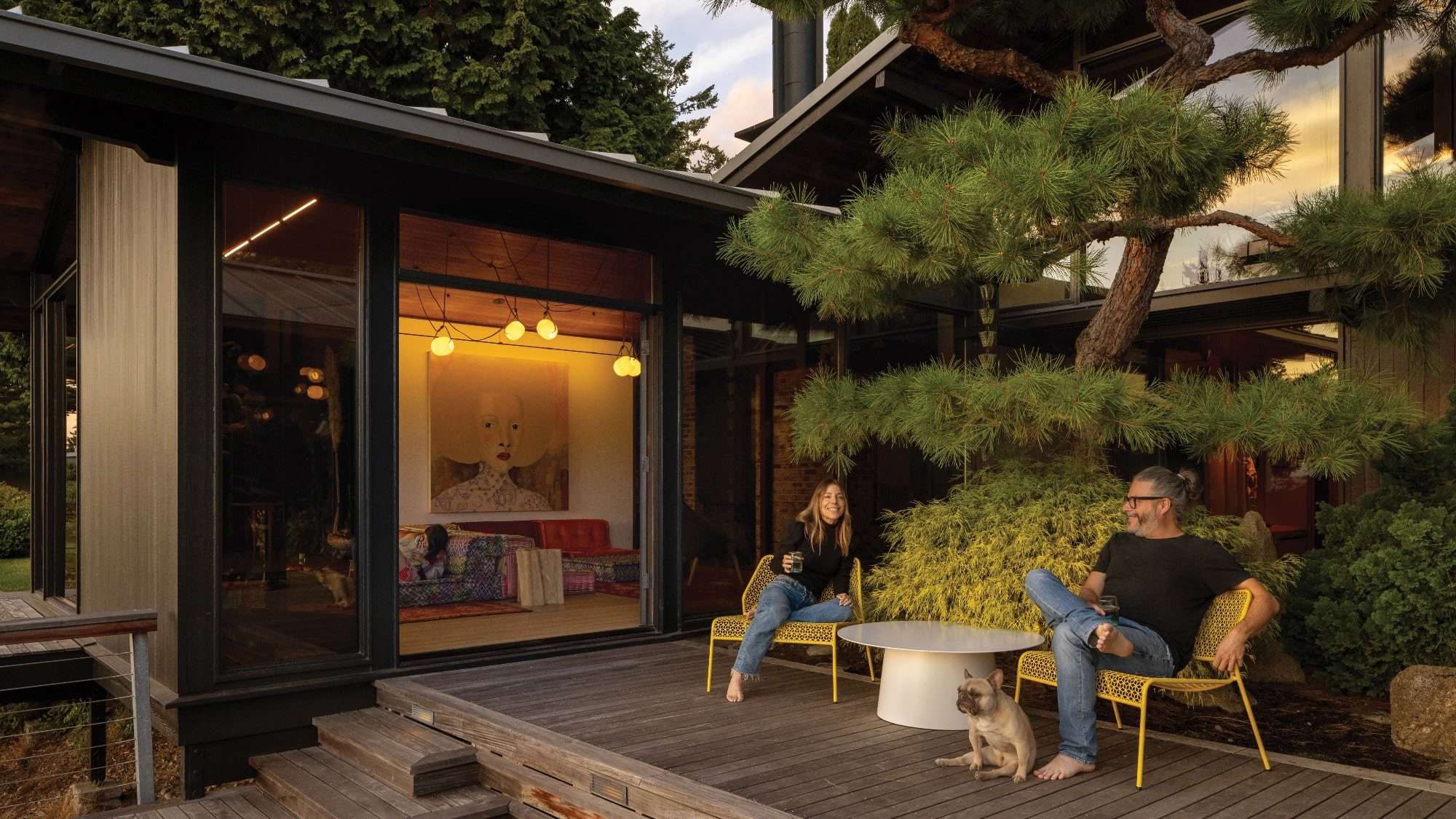 A man and woman sit on yellow chairs beside a small table on a wooden deck, with a dog at their feet. The deck, recently enhanced through remodeling, is adjacent to a modern house with large windows and lush greenery.